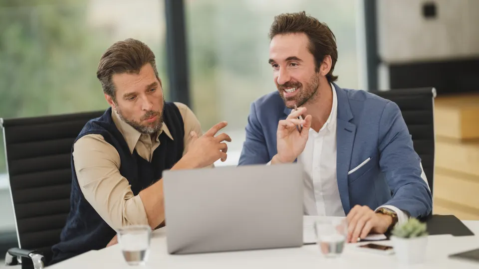 Two men in blazers talking over a laptop at a conference table.