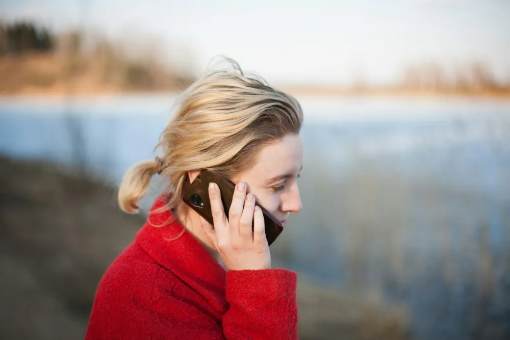 A woman in a red sweater outdoors by the water, holding a phone to her ear and looking down while she listens.
